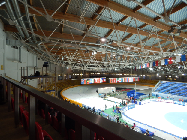 Große Indoor-Eisfläche mit zahlreichen Menschen, die Schlittschuh laufen, umgeben von Sitzplätzen, Geländern, Brettern, Beleuchtungskörpern und konstruktiven Elementen, mit einer Wand im Hintergrund.