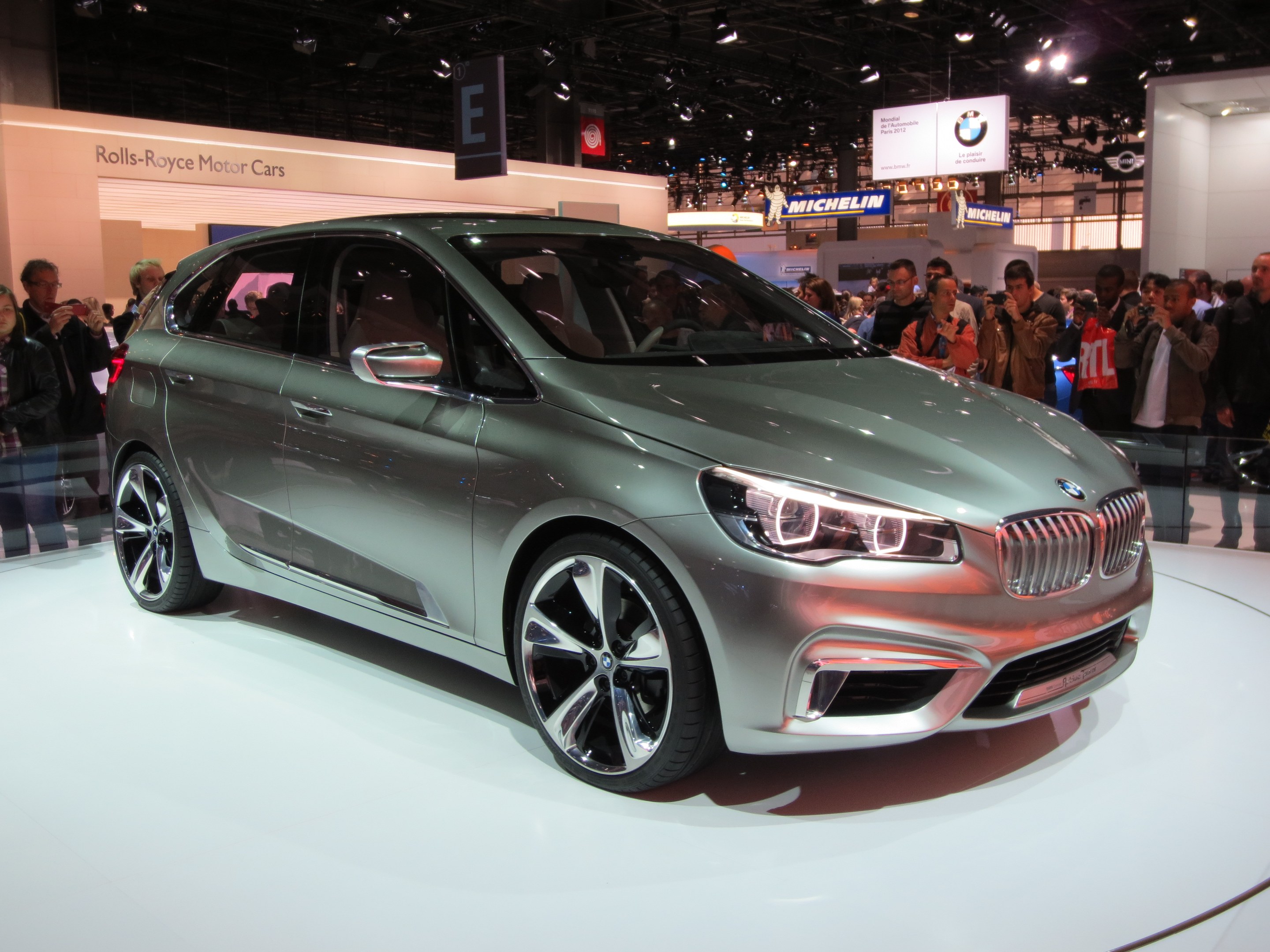 Ein BMW 2er Active Tourer Concept-Auto auf der Frankfurter Motor Show, umgeben von Menschen, die es unter Deckenbeleuchtung fotografieren, mit Texttafeln im Hintergrund.