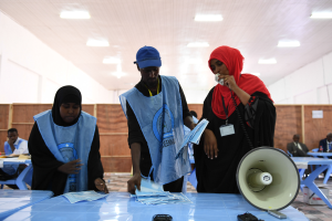 Eine Gruppe von Menschen steht um einen Tisch mit einem Megafon, Papieren und anderen Gegenständen, während andere im Hintergrund auf Stühlen sitzen oder stehen, unter einer Decke mit Lampen, was auf eine Wahllokal hindeutet.