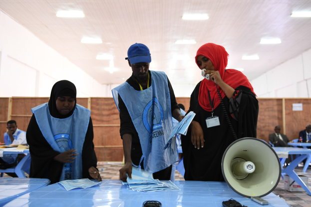 Eine Gruppe von Menschen steht um einen Tisch mit einem Megafon, Papieren und anderen Gegenständen, während andere im Hintergrund auf Stühlen sitzen oder stehen, unter einer Decke mit Lampen, was auf eine Wahllokal hindeutet.