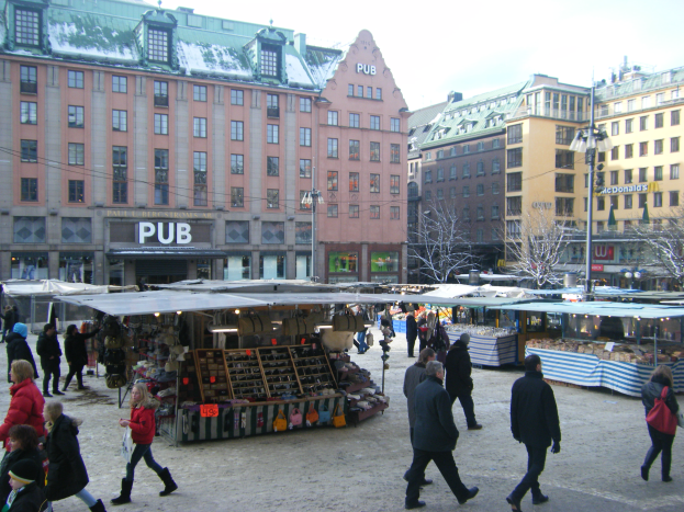 Ein lebendiger Weihnachtsmarkt in Stockholm, Schweden, mit Menschen, die herumlaufen, Ständen voller Gegenstände, Gebäuden, Laternen, Bäumen und einem klaren blauen Himmel.