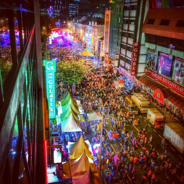 Eine volle Jahrmarktszene mit Zelten, Geschäften, Gebäuden und Bannern, mit hell erleuchteter Fläche im Hintergrund.