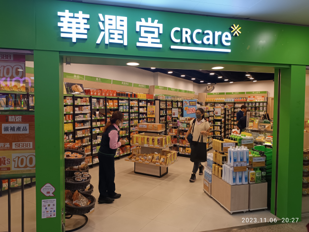 Eine Gruppe von Menschen steht vor einem Geschäft mit einer Tafel, auf der "crcare" steht, mit Regalen voller Lebensmittel, Flaschen und anderen Gegenständen im Inneren und einer Tafel mit Text auf der linken Seite.