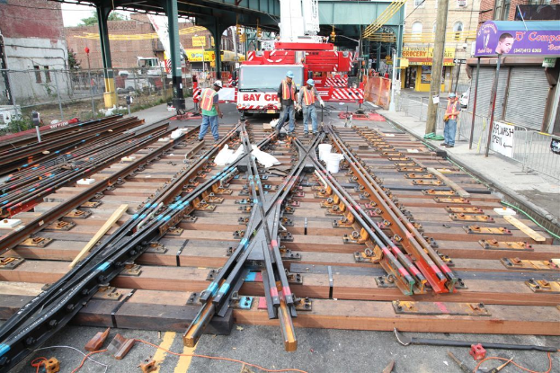 Arbeiter in Jacken und Mützen bauen Bahnschienen, mit einem Fahrzeug voller Materialien und einer Hütte mit Pfählen im Hintergrund und einem Geschäft auf der anderen Seite.