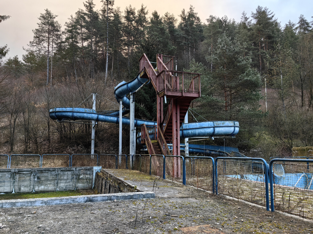 Ein verlassenes Wasserpark mit einer blauen Wasserrutsche, Geländern, Treppen und einer Wand, umgeben von Bäumen und einem klaren blauen Himmel, mit Text am unteren Bildrand.
