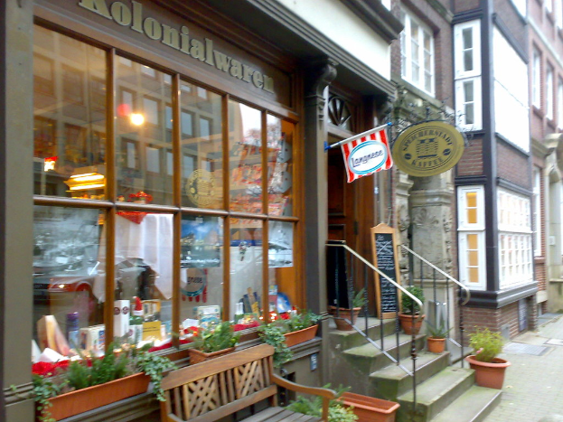 Eine Szene mit Gebäuden, Texttafeln, Pflanzen, einem Fenster mit Gegenständen dahinter, Text über dem Fenster, Treppen mit Geländern und einer leeren Bank.