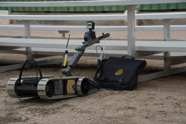 Ein Roboter sitzt auf einem Schmutzfeld neben einer Bank, mit einer Kamera und einer Tasche auf dem Boden, vor einem Zaun und einer Wand, was eine Robotik-Wettbewerbs-Situation suggeriert.