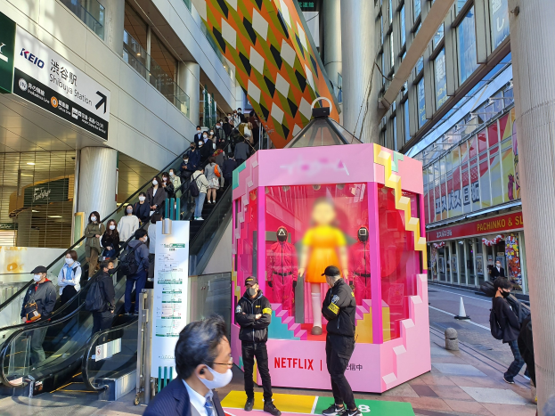 Eine Gruppe von Menschen steht vor einem Gebäude mit Glaswänden, Säulen und Texttafeln, einige tragen Masken. Eine Rolltreppe mit Menschen ist zu sehen, und eine pinkfarbene Box in der Mitte zeigt "Netflix's new Netflix booth in Tokyo" mit Schaufensterpuppen darin.