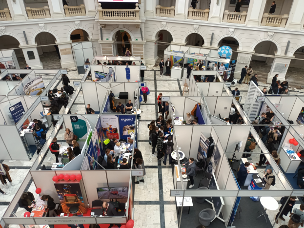 Eine große Gruppe von Menschen auf einer internationalen Fachmesse mit Ständen, Bannern, Tischen, Stühlen, Ballons und einem Gebäude mit Bögen, Säulen, Geländern und einem Bildschirm im Hintergrund.