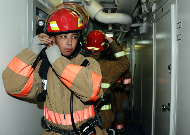 Feuerwehrleute in Uniform stehen in einem Raum mit Rohren und Gegenständen im Hintergrund, wahrscheinlich während eines Trainings.
