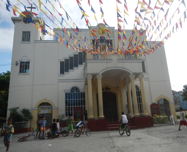 Eine Gruppe von Menschen, die vor einer weißen Kirche mit architektonischen Details, Pflanzen, einem Zaun, einer Fahne, Bäumen und einem bewölkten Himmel Fahrrad fahren, mit bunten Fahnen, die an einem Seil befestigt sind.
