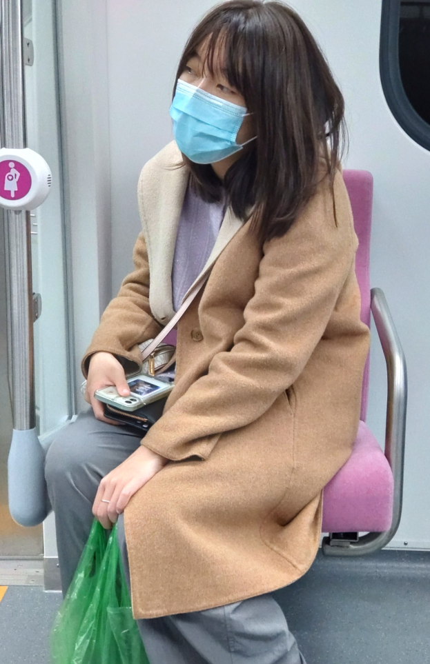 Eine Frau mit Mund-Nasen-Schutz sitzt in einem Zug, hält ein Mobiltelefon und eine Tasche und hat eine Tür links von ihr.