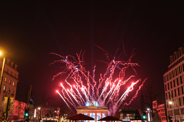 Eine belebte Stadtstraße in Berlin am Silvesterabend, voller Menschen, Fahrzeuge und Gebäude, beleuchtet von Feuerwerk und Gebäudelichtern, die eine festliche Atmosphäre schaffen.