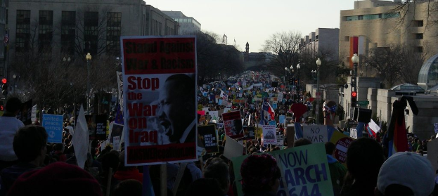 Menschen mit Plakaten auf einer Straße, mit Gebäuden, Bäumen, Laternenmasten und Himmel im Hintergrund.