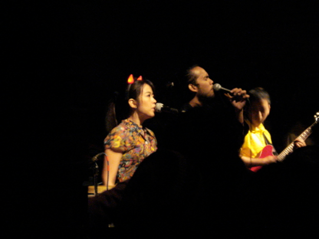 Eine Frau und ein Mann singen in ein Mikrofon, während eine Person eine Gitarre spielt.