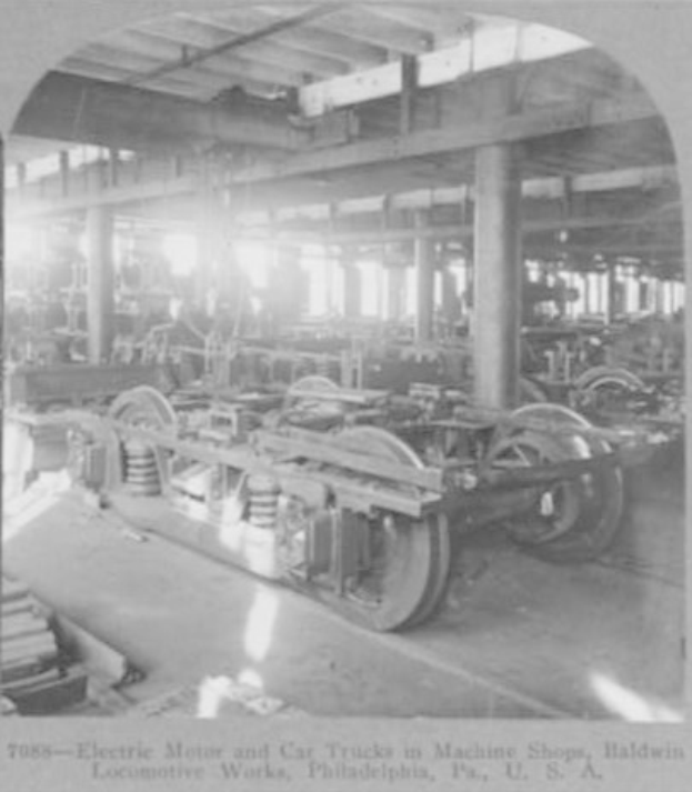 Schwarzes und weißes Foto eines Elektromotors und eines Lkw in einer Fabrik mit Maschinen, Säulen und einem Dach, mit Text unten.