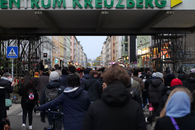 Eine belebte Straße in Berlin, Deutschland, mit einer Menge Menschen, die an dem "Entrum Kreuzberg"-Gebäude vorbeigehen und Fahrräder fahren, unter einem klaren blauen Himmel.