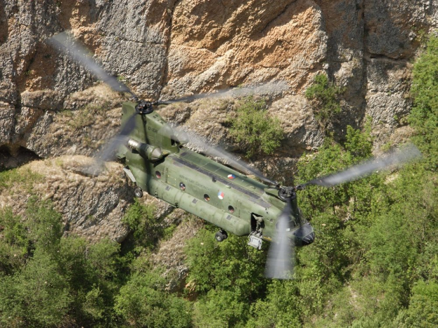 Ein Milit√§rhelikopter √ľberfliegt eine felsige Klippe mit B√§umen im Vordergrund.