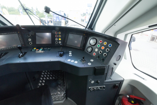 Ein Zugcockpit mit einem Feuerlöscher im Vordergrund, ein Glasfenster auf der rechten Seite und eine Außenszene mit Menschen, Fahrzeugen, Bäumen und Masten im Hintergrund.