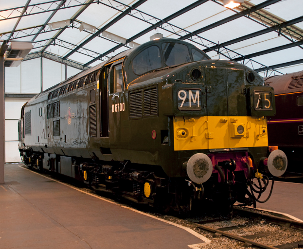 Ein Zug steht auf den Schienen neben einem Bahnsteig mit einem an der Decke befestigten Pfahl, der Metallstangen mit Lampen trägt.