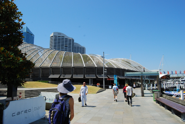 Menschen, die auf einem Gehweg mit Mülltonnen, Bänken, Holzlatten und Metallstangen auf der rechten Seite, Pfählen, Bäumen und einem Metallstangenzaun mit Bannern auf der linken Seite und Gebäuden im Hintergrund gehen.