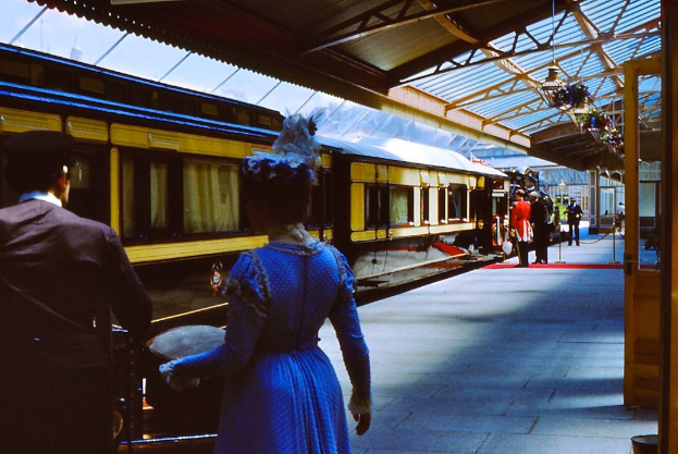 Frau in blauem Kleid neben einem Zug an einem Bahnhof mit einigen Leuten auf dem Bahnsteig.