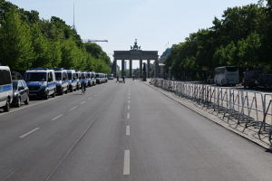 Lange Reihe von Polizeiwagen, die vor dem Brandenburger Tor auf der Straße geparkt sind, mit Fahrradfahrern und Füssgängern, Barrieren, Bäumen und dem Torbogen mit Statuen im Hintergrund.