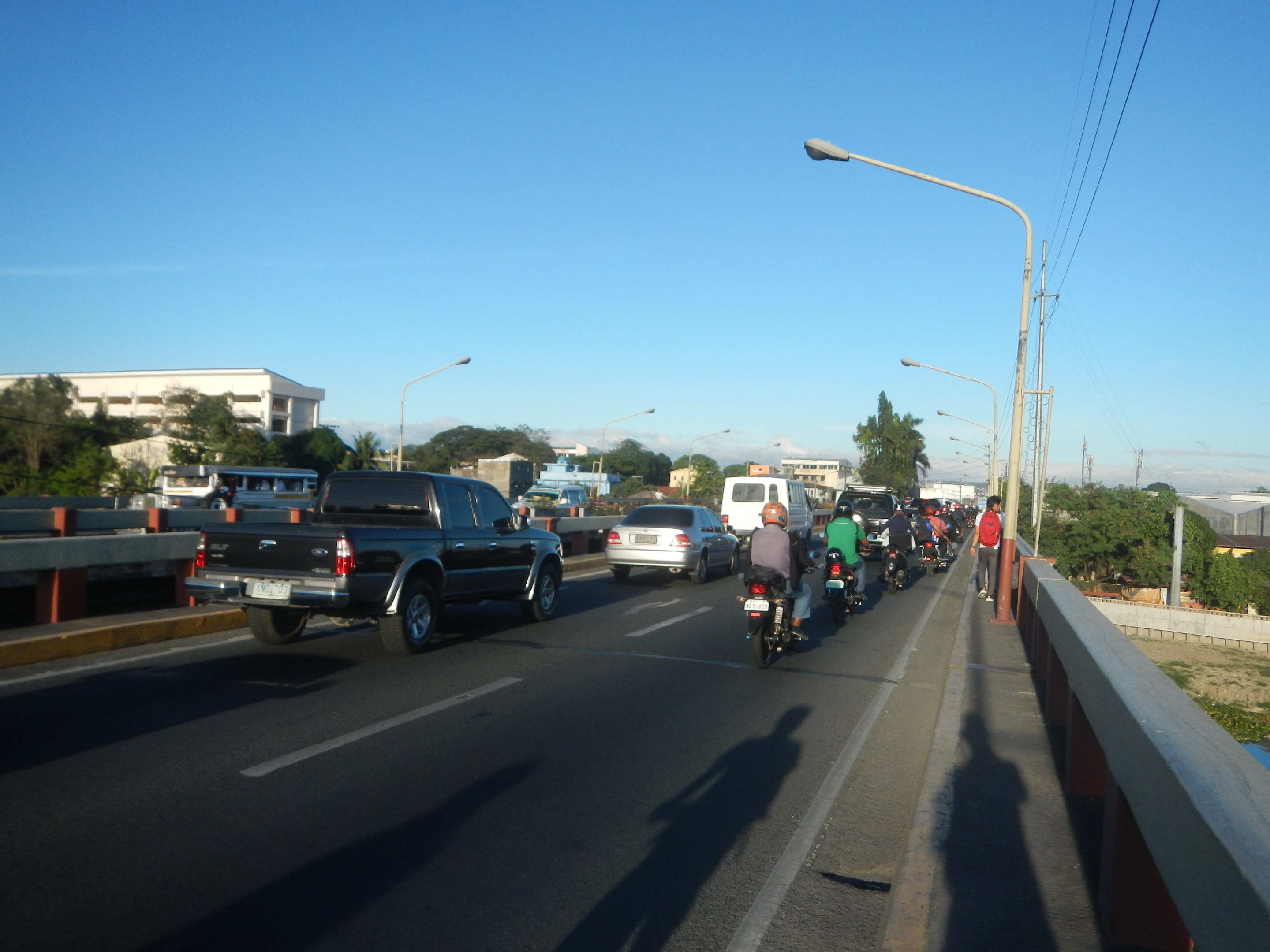 Eine Gruppe von Motorradfahrern überquert eine Brücke mit einem Pick-up-Truck im Vordergrund, unter einem klaren blauen Himmel mit Bäumen und Gebäuden im Hintergrund.