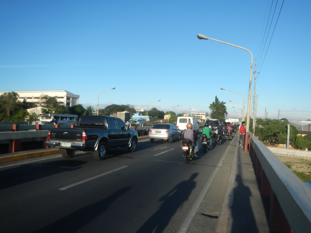 Eine Gruppe von Motorradfahrern überquert eine Brücke mit einem Pick-up-Truck im Vordergrund, unter einem klaren blauen Himmel mit Bäumen und Gebäuden im Hintergrund.