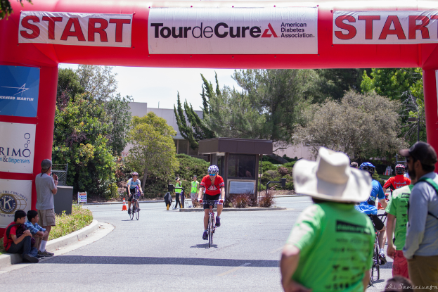 Eine belebte Straßenszene mit Menschen, die Fahrräder fahren, einige auf einem Fußweg sitzen und andere auf der Straße stehen, mit einer Hütte und umgebenden Pflanzen und Bäumen im Hintergrund.