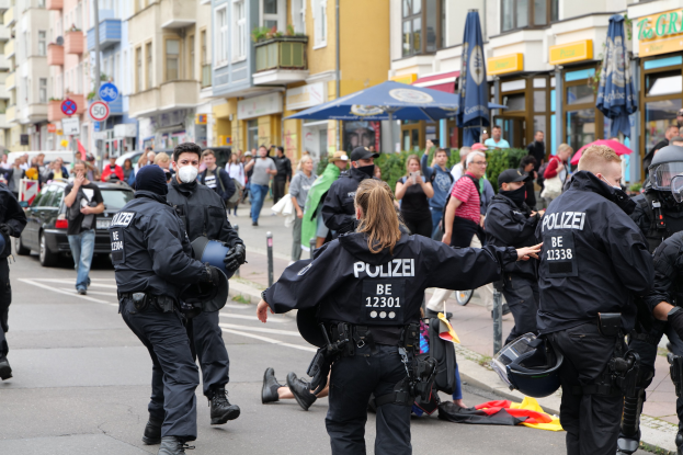 Eine Gruppe von Polizeibeamten geht eine Straße entlang neben einer Menschenmenge, wobei einige Helme und Handschuhe tragen oder Schirme halten, mit Fahrzeugen, Schildern, Pflanzen und Gebäuden im Hintergrund.