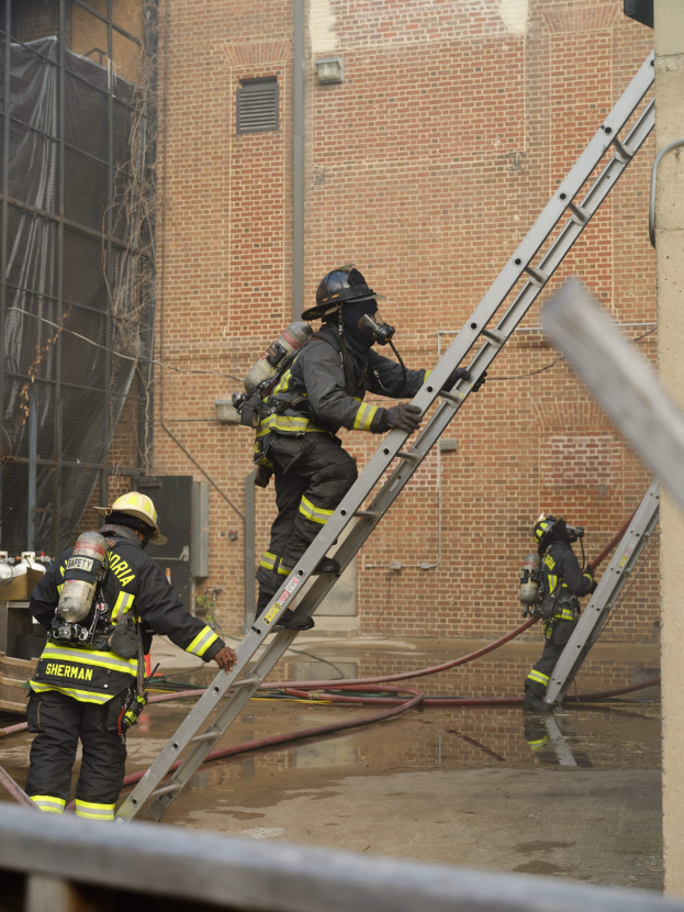 Feuerwehrleute in Helmen und Ausr"ustung klettern an einer Leiter vor einem Backsteingeb"ude mit Rohren auf dem Boden und einer Metallstange unten, mit einem anderen Geb"ude und einem Netz im Hintergrund.