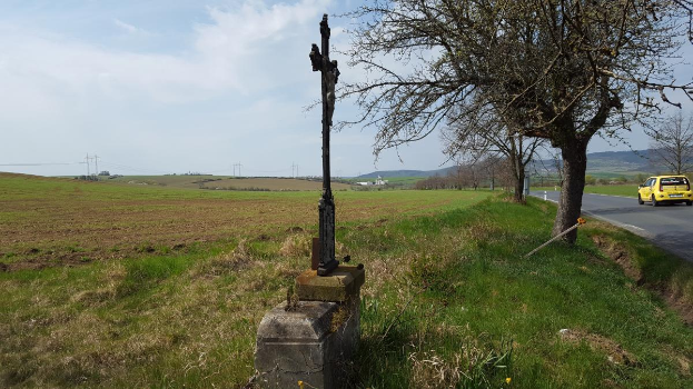 Ein gelbes Auto fährt auf einer Straße neben einem Baum, mit einem Kreuz am Straßenrand, Hügeln und einem bewölkten Himmel im Hintergrund und Gras und Blumen, die den Boden bedecken.