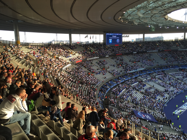 Eine große Menschenmenge sitzt im Allianz Arena Stadion in München, Deutschland, und schaut ein Fußballspiel. Auf der rechten Seite befindet sich eine Bühne, Fahnen, Stangen und ein Bildschirm im Hintergrund, und der Himmel ist oben sichtbar.