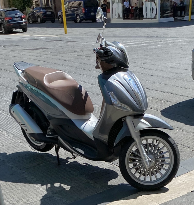 Ein Motorroller mit einem Helm darauf auf einer Straße geparkt, mit Gebäuden, Bäumen, Pfählen, Menschen, Stühlen, Tischen und anderen Objekten im Hintergrund.