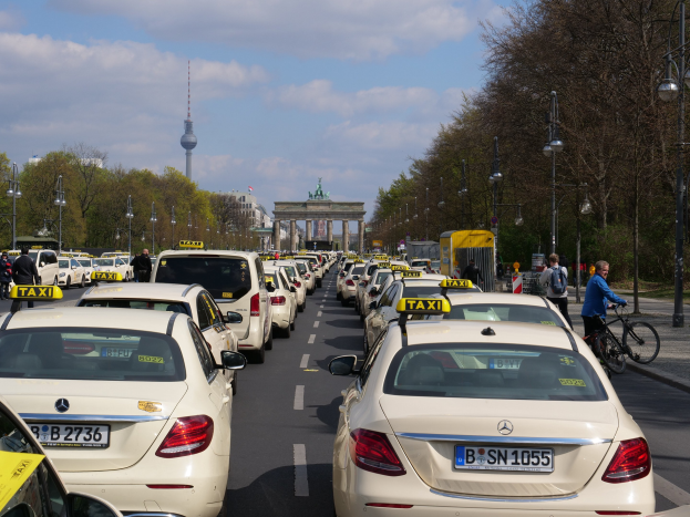 Eine lange Reihe von Taxis, die an einer belebten Straße in Berlin, Deutschland, geparkt sind, mit Fahrradfahrern und Fußgängern auf dem Gehweg, flankiert von Laternen und Bäumen und Gebäuden, einem Bogen und einem Turm im Hintergrund unter einem bewölkten Himmel.