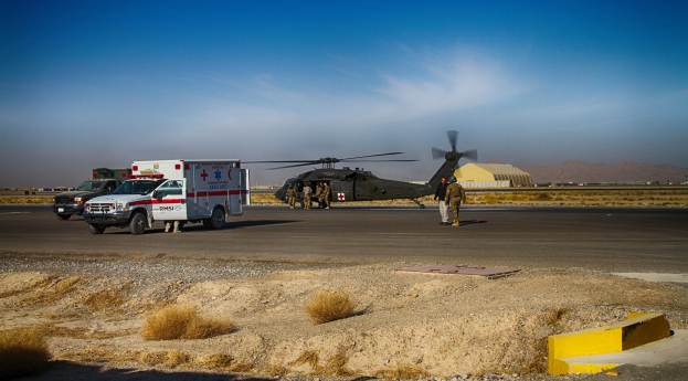 Ein Hubschrauber neben einem Rettungswagen auf einer grasbewachsenen Straße geparkt, mit Menschen in der Nähe, Bergen im Hintergrund und einem klaren blauen Himmel.