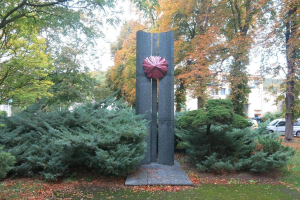 Große Metallskulptur inmitten eines Parks mit Bäumen, Gras und welken Blättern, umgeben von Gebäuden und einem klaren blauen Himmel, Teil der Kunst der Stadt Berlin.