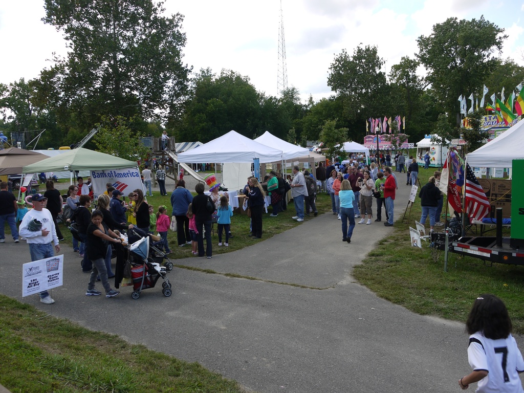 Eine Gruppe von Menschen, Stühle, Flaggen, Kinderstühle, Zelte und eine Tafel sind auf einer grünen Fläche mit Bäumen im Hintergrund aufgebaut.