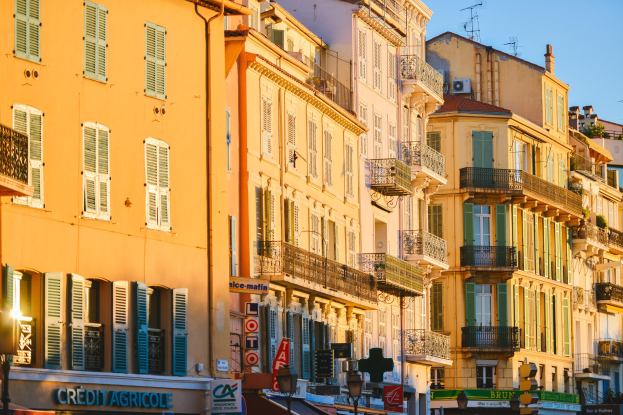 Eine Stadtstraße mit hohen Gebäuden, die Fenster, Balkone und Schilder zeigen, gesäumt von Bäumen unter einem klaren blauen Himmel.