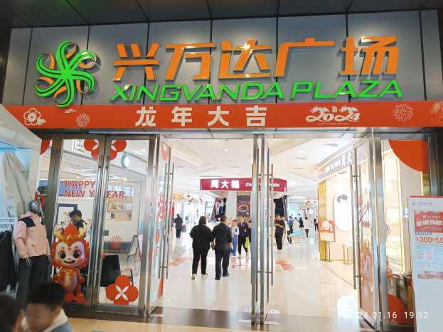 Der Eingang zum Xingvanda Plaza Einkaufszentrum, mit Menschen, die hindurchgehen, Kleider an Bügeln links, ein Textschild rechts, Deckenlampen darüber und zwei Kinder unten.