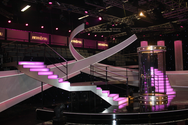 Eine Bühne mit einer Wendeltreppe, Geländern, Stufen, Beleuchtungsgeräten, Stangen, Texttafeln, Säulen und anderen Requisiten vor einem dunklen Hintergrund.