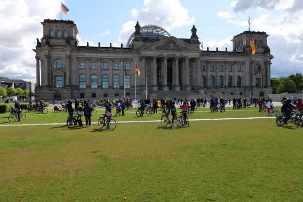 Eine Gruppe von Menschen auf Fahrrädern vor dem Reichstaggebäude in Berlin, Deutschland, das mit Fenstern, Säulen und Fahnen mit Stangen geschmückt ist, umgeben von Bäumen und Pflanzen, mit einem bewölkten Himmel und grasbewachsenem Boden.