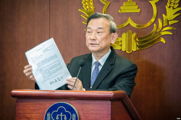 Ein Mann im Anzug und Krawatte steht an einem Rednerpult, hält ein Blatt Papier in der Hand und spricht in ein Mikrofon, mit einem Logo im Hintergrund, das seine Rolle als neu ernannter Chief Justice der Volksrepublik China anzeigt.