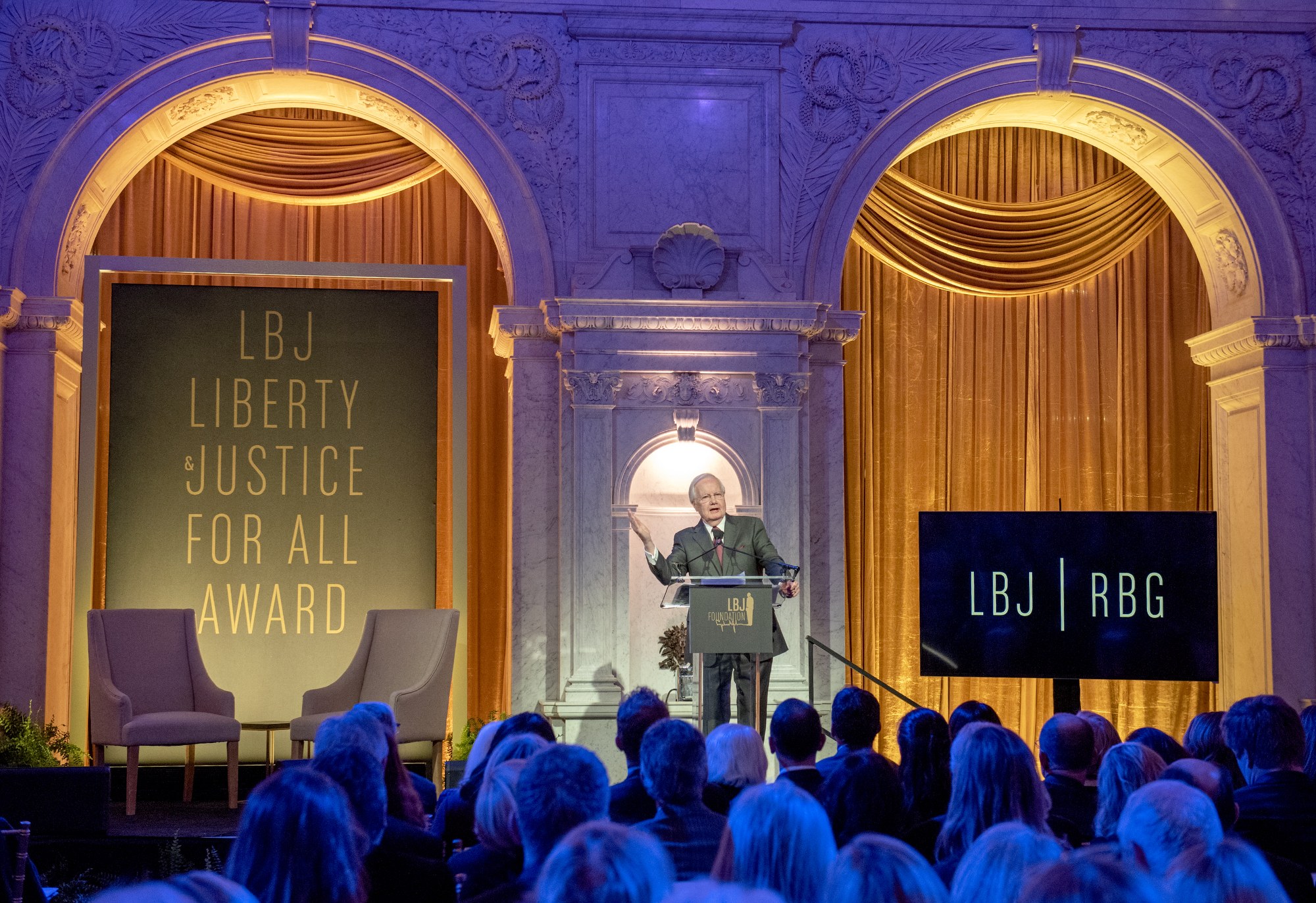 Ein Mann steht an einem Podium mit einem Mikrofon und spricht zu einer sitzenden Menge in einem Raum mit Stühlen, Pflanzen, Vorhängen und einer Tafel mit Text dahinter.