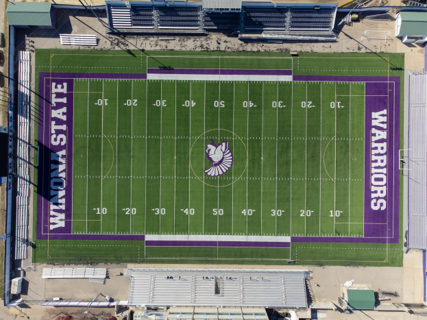 Eine Luftaufnahme eines Footballfeldes mit violetten und weißen Markierungen, umgeben von Gebäuden, Fahrzeugen und Masten, die das Heimfeld der Footballmannschaft der Minnesota State University zeigt.