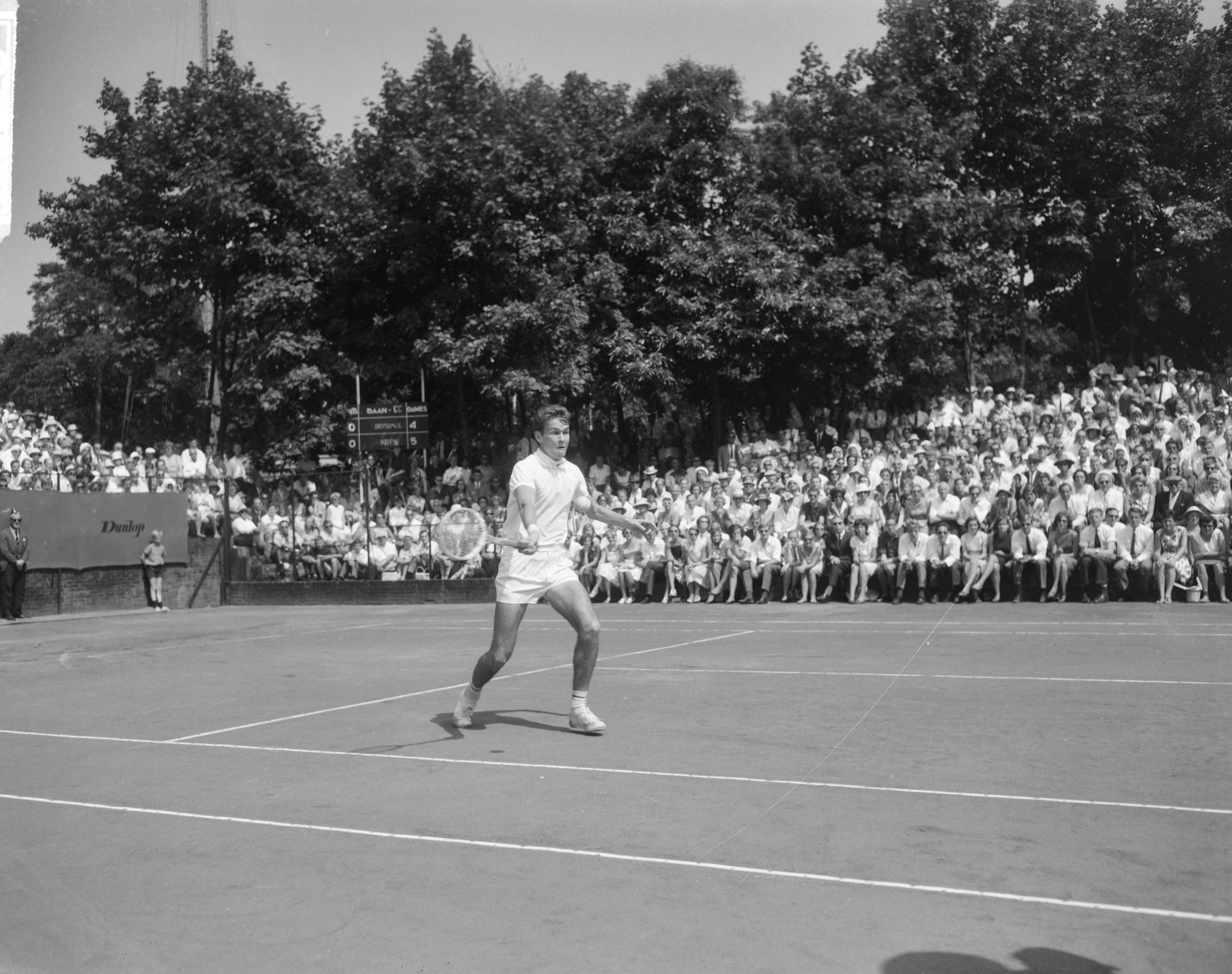 Ein Mann spielt Tennis auf einem Platz mit einer Menge von Menschen, Bäumen und einem Turm im Hintergrund unter einem klaren Himmel.