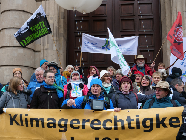 Eine Gruppe von Menschen steht vor einem Gebäude mit Säulen und einer Tür, hält ein Transparent mit Text, Fahnen und Ballons und nimmt an einem Klimastreik in Deutschland teil.