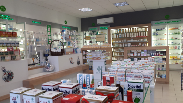 Ein Apothekeninneres mit verschiedenen Medikamenten auf Tischen, einem Monitor und einem Korb an den Seiten, Regalen im Hintergrund und Deckenlampen.