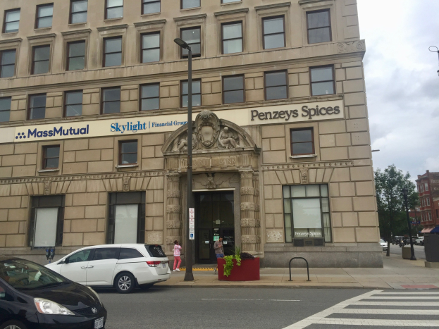 Großes Gebäude mit parkenden Autos davor, Straßenlaternen, fahrenden Fahrzeugen, Fußgängern auf dem Gehweg, Grünfläche und einem bewölkten Himmel, identifiziert als Hauptsitz der Massachusetts Mutual Financial Group.